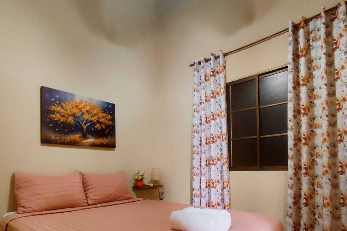 A bedroom with a bed, a nightstand, a painting, and a window with curtains.