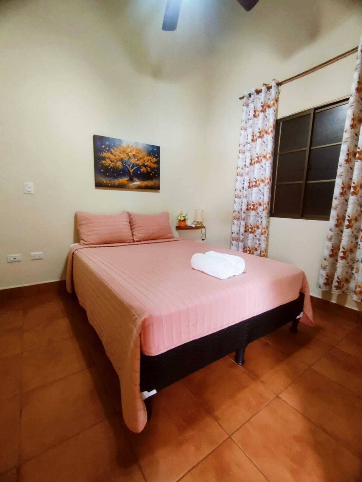 A bedroom with a bed, a nightstand, a painting, and a window with curtains.