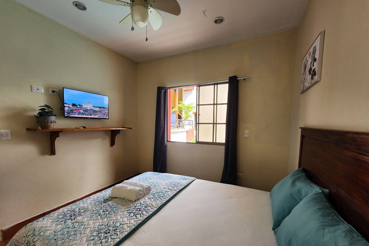 A bedroom with a bed, a TV on the wall, a window with curtains, and a ceiling fan.