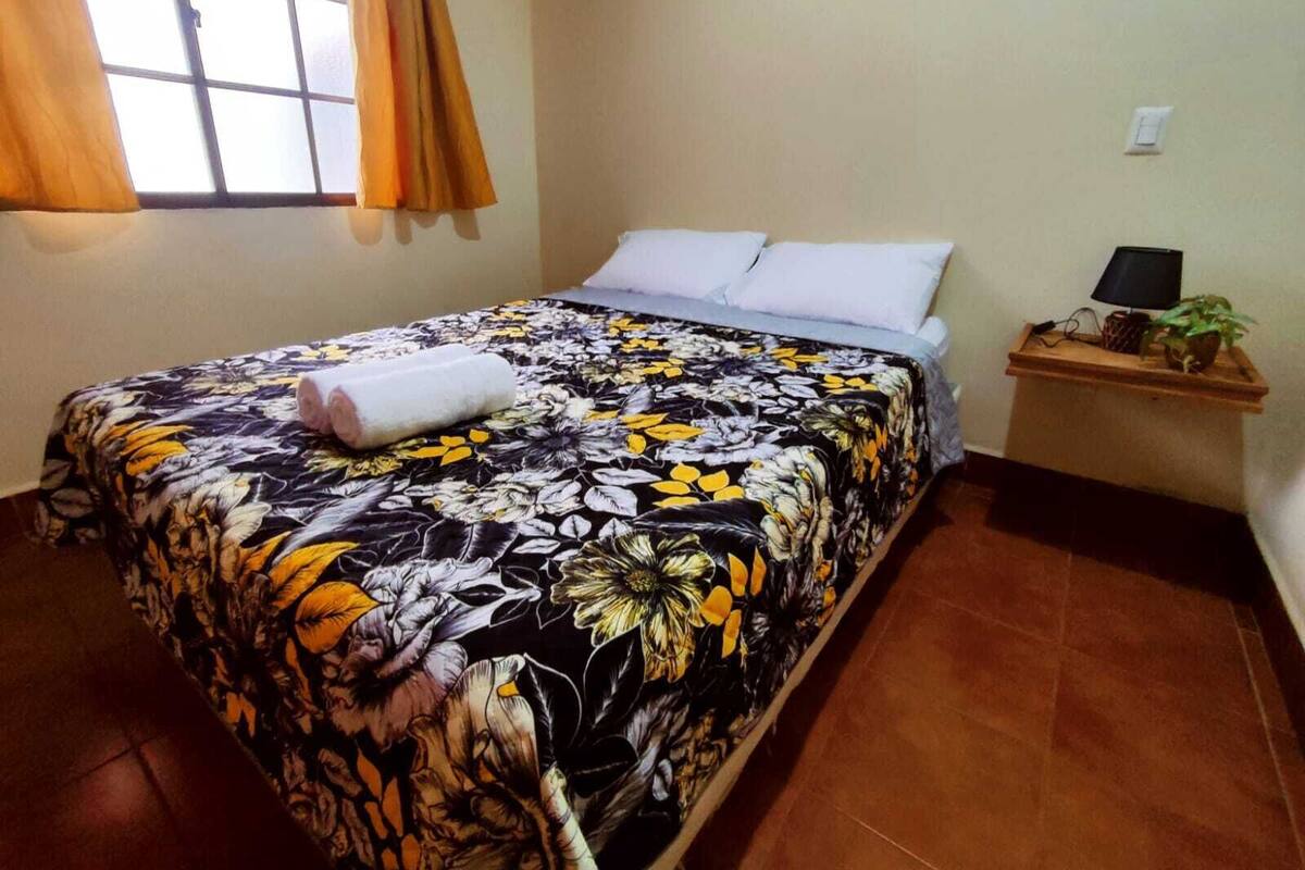 A neatly made bed with a floral bedspread, a rolled towel, and a bedside table with a lamp and a plant.
