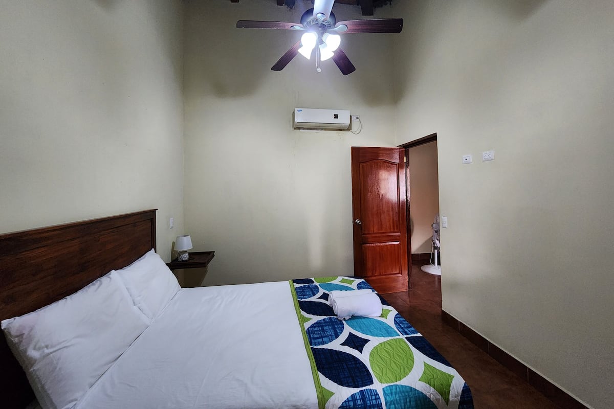 A hotel room with a bed, a nightstand, a ceiling fan, a door, and an air conditioning unit.