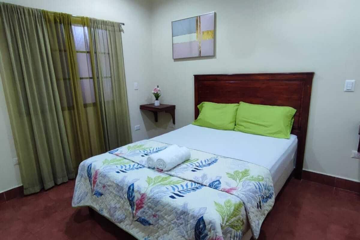 A bedroom with a bed, a ceiling fan, a window with curtains, and a small bedside table with a vase.