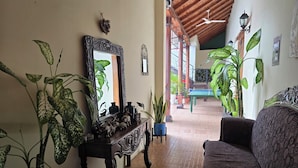 A living room with a sofa, a coffee table, and a large mirror. There are potted plants and a wooden bench. The room has a tiled floor and a ceiling fan. - Casona Macondo (Granada)