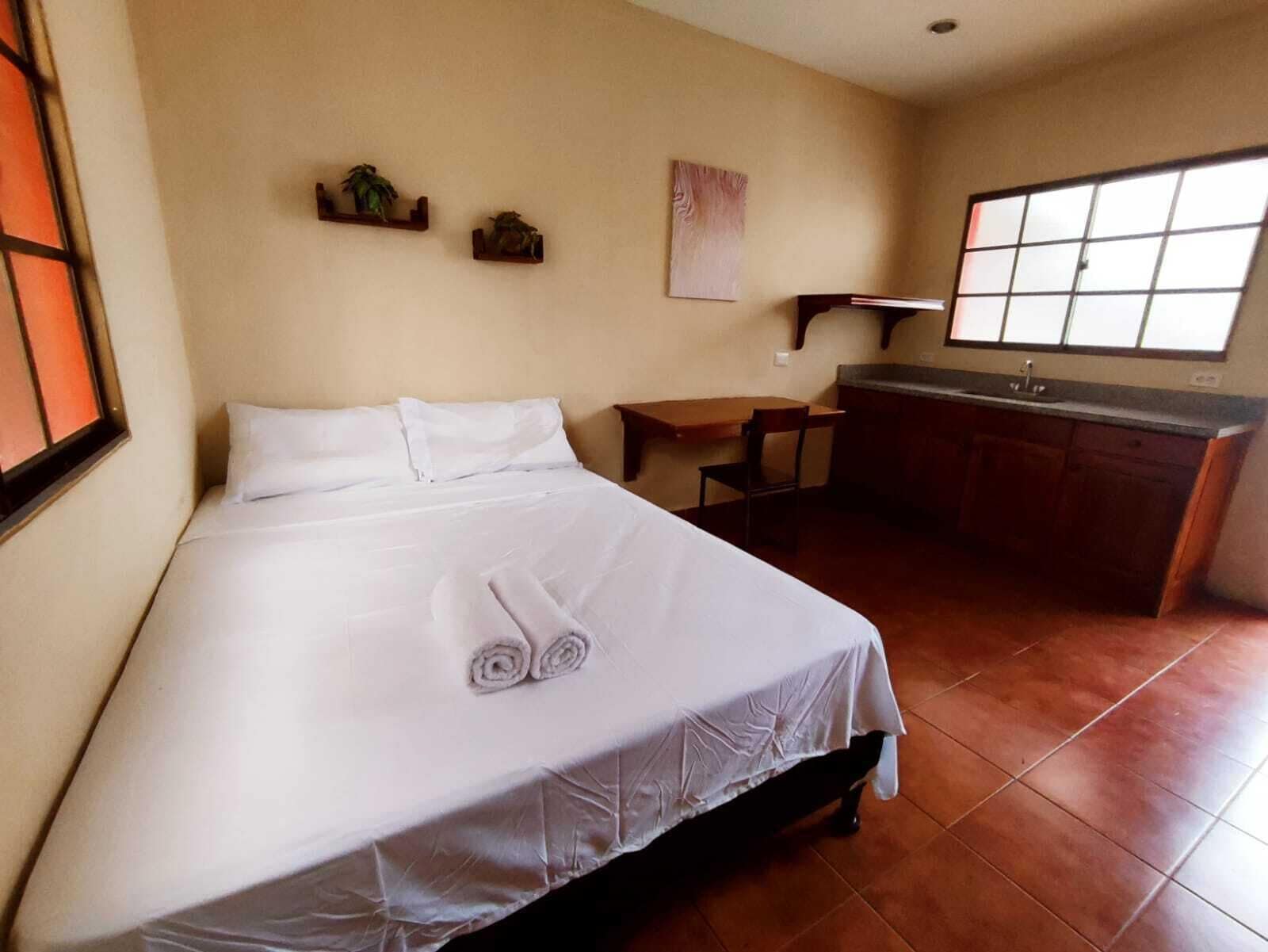 Une chambre avec un lit, une petite table, une chaise, une fenêtre et un coin cuisine.
