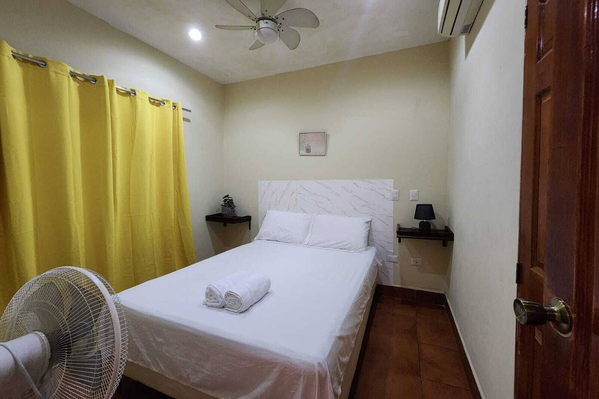A single bed with a white headboard, a bedside table with a lamp, a ceiling fan, and a yellow curtain.