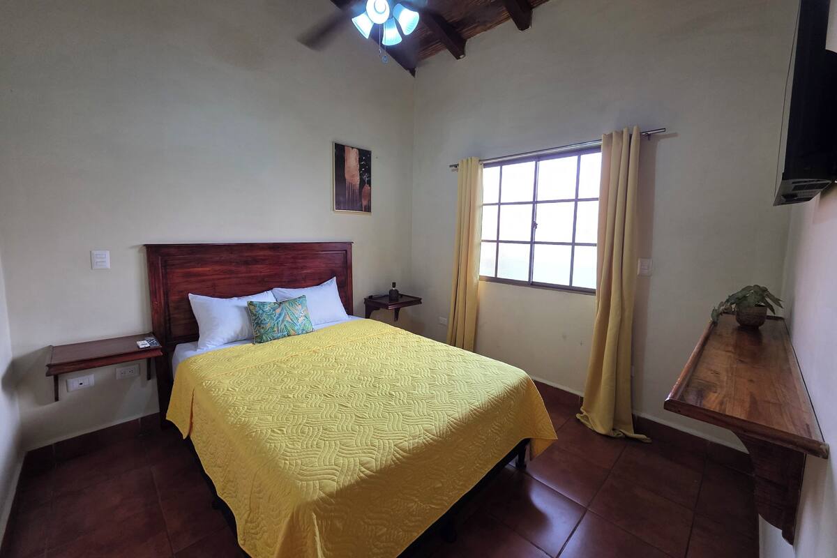 A bedroom with a wooden bed, a ceiling fan, a window with curtains, and a small table with a plant.