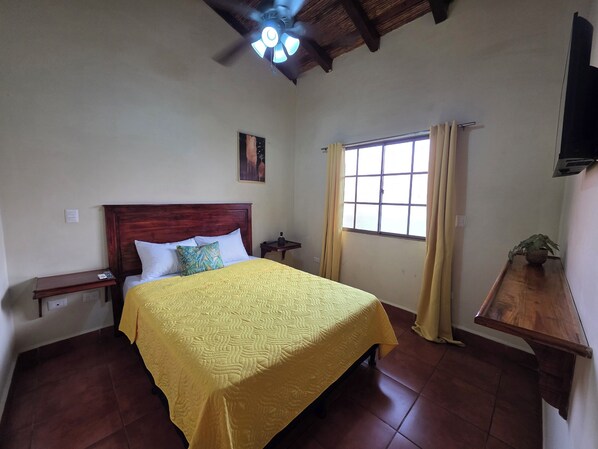 A bedroom with a wooden bed, a ceiling fan, a window with curtains, and a small table with a plant. - Casona Macondo (Granada)