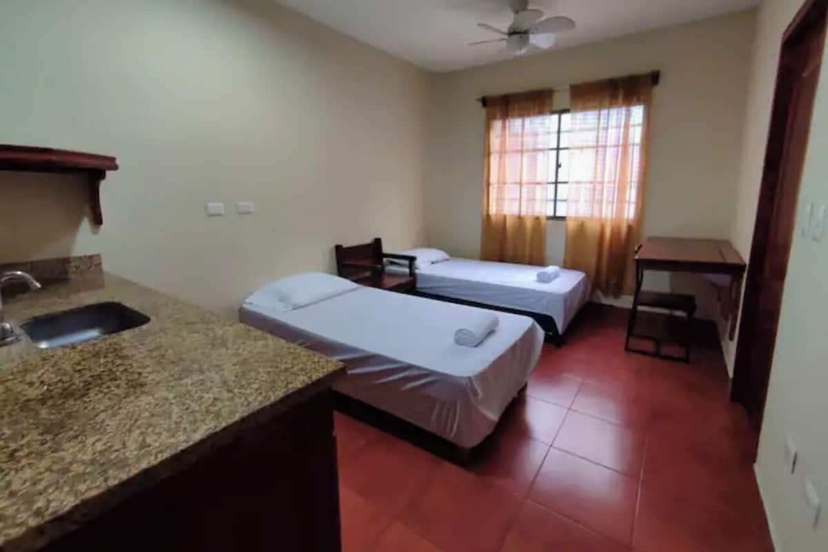 A room with two beds, a small kitchen area with a sink, a ceiling fan, and a window with curtains.