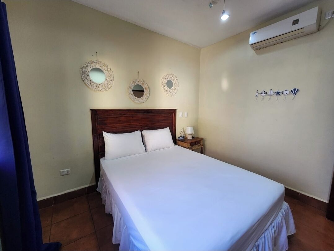 A bedroom with a large bed, a wooden headboard, two wall-mounted mirrors, a nightstand with a lamp, and a wall-mounted air conditioner.