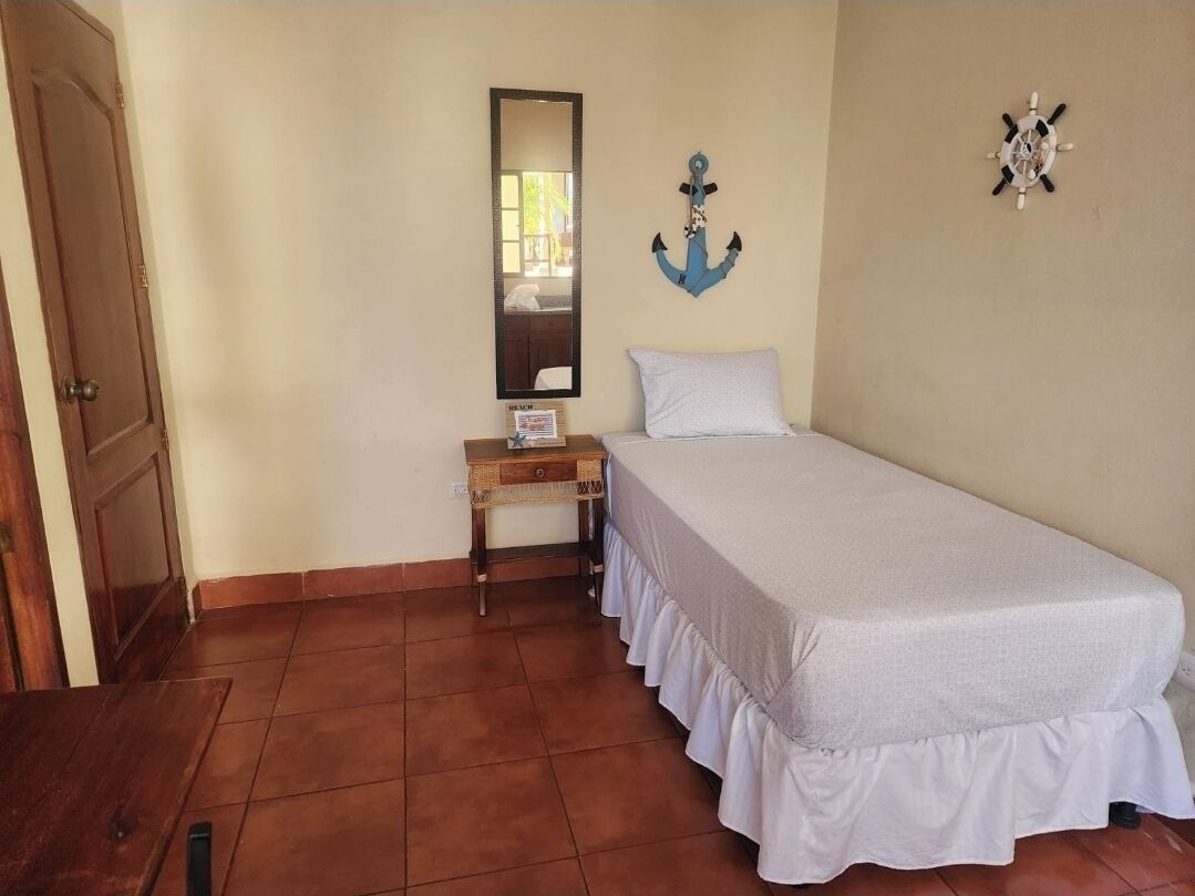 Una cama individual con cubrecama blanco, una mesita de noche de madera con lámpara, un espejo fijado a la pared y una decoración de ancla azul.