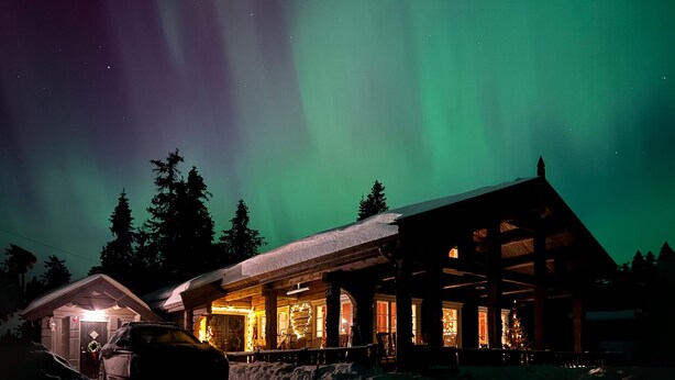 Una cabaña de troncos con techo cubierto de nieve e interior iluminado, rodeada de árboles bajo un cielo nocturno con la aurora boreal.