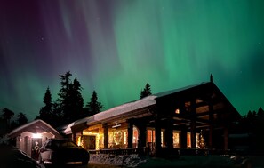A log cabin with a snow-covered roof and a lit interior, surrounded by trees under a night sky with the aurora borealis.