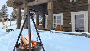 Una cabaña de madera con porche cubierto, una fogata y un paisaje nevado.