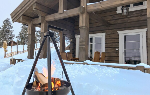 A wooden cabin with a covered porch, a fire pit, and snow-covered surroundings.