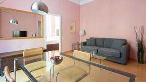A modern living room with a glass dining table, two wicker chairs, a grey sofa, a large mirror, and a potted plant.