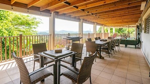 Een overdekt terras met een eethoek, rieten stoelen en tafels, een barbecue en uitzicht op het omliggende landschap.