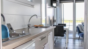 Et moderne køkken med vask i rustfrit stål, elkedel og brødrister. Spiseområde med bord og stole er synligt igennem en glasdør.