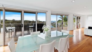 A modern interior with a glass dining table, white chairs, and a balcony with outdoor seating. - Three Sisters | Ocean Views & Walk to Beach (Long Beach)