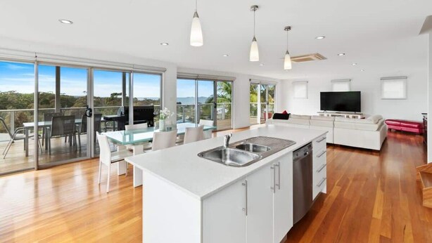 A modern kitchen with a large island, a dining area with glass doors, and a living room with a flat-screen TV. - Three Sisters | Ocean Views & Walk to Beach (Long Beach)
