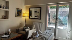 Een woonkamer met een bank, een salontafel, een lamp en een radiator. Een deur leidt naar een balkon met uitzicht op een bakstenen muur en groen.