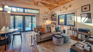 A spacious living room with a wooden ceiling, a leather sofa, a dining area with a table and chairs, and large windows offering views of the outdoors. - Private Forest Retreat • Spa Bath • Near the Coast • Central Midcoast Hub (Searsport)