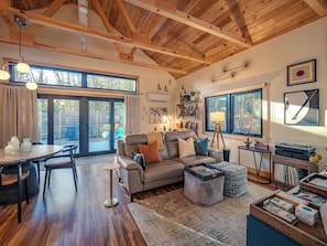 Un salón espacioso con techo de madera, sofá de cuero, comedor con mesa y sillas, y ventanas grandes que ofrecen vistas al exterior.