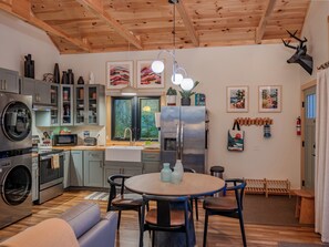 A modern kitchen with stainless steel appliances, a dining area with a round table, and a wooden ceiling. - Private Forest Retreat • Spa Bath • Near the Coast • Central Midcoast Hub (Searsport)
