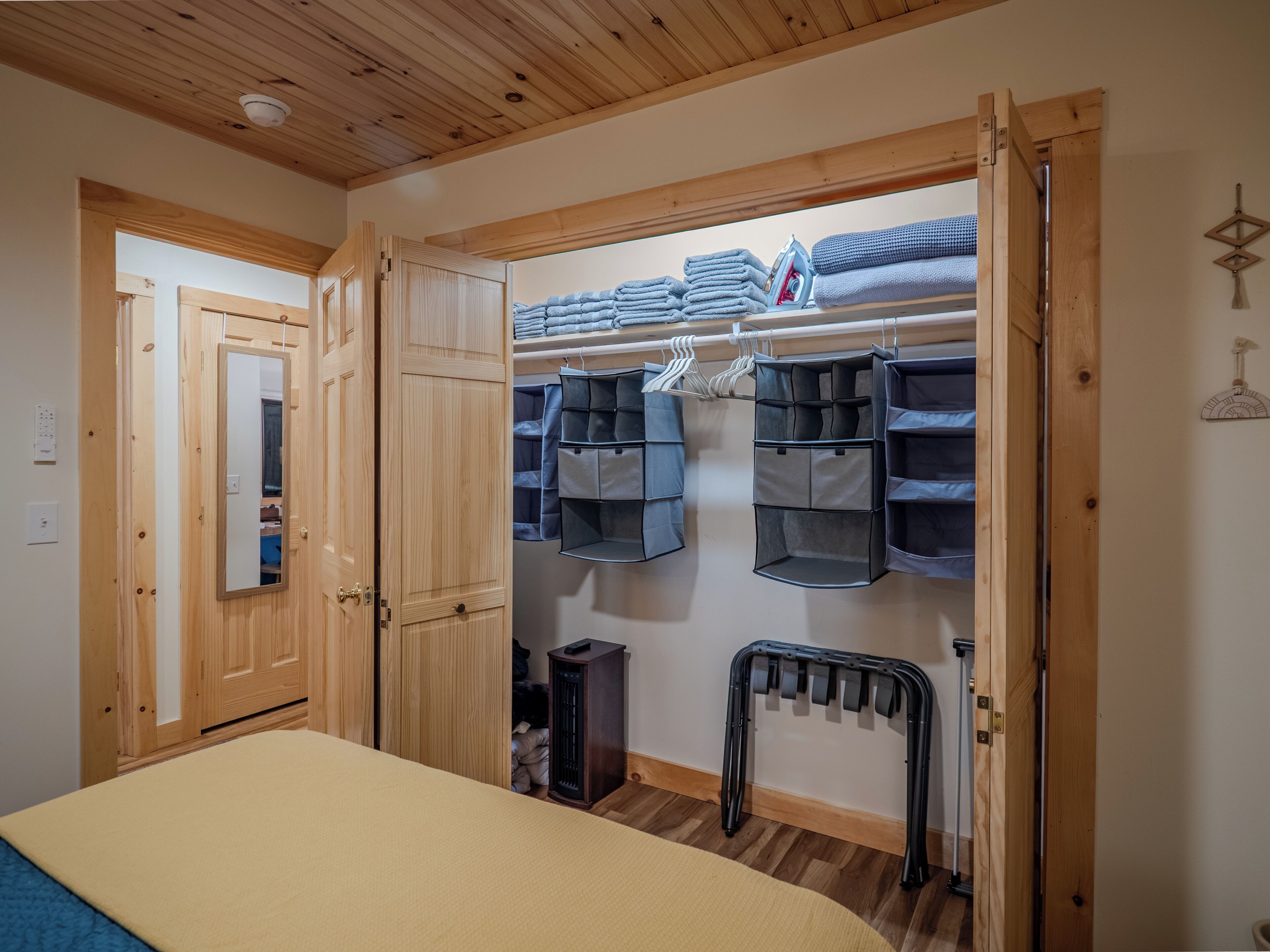Une chambre avec un plafond en bois, un lit recouvert d’une couverture beige, une armoire en bois, un placard avec des étagères et des organisateurs suspendus, et un radiateur.