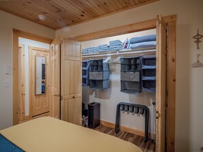 A bedroom with a wooden ceiling, a bed with a beige blanket, a wooden wardrobe, a closet with shelves and hanging organizers, and a radiator.