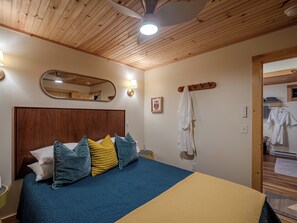 Habitación de hotel con techo de madera, cama con ropa de cama azul y amarilla, mesita de noche, espejo y perchero en la pared.