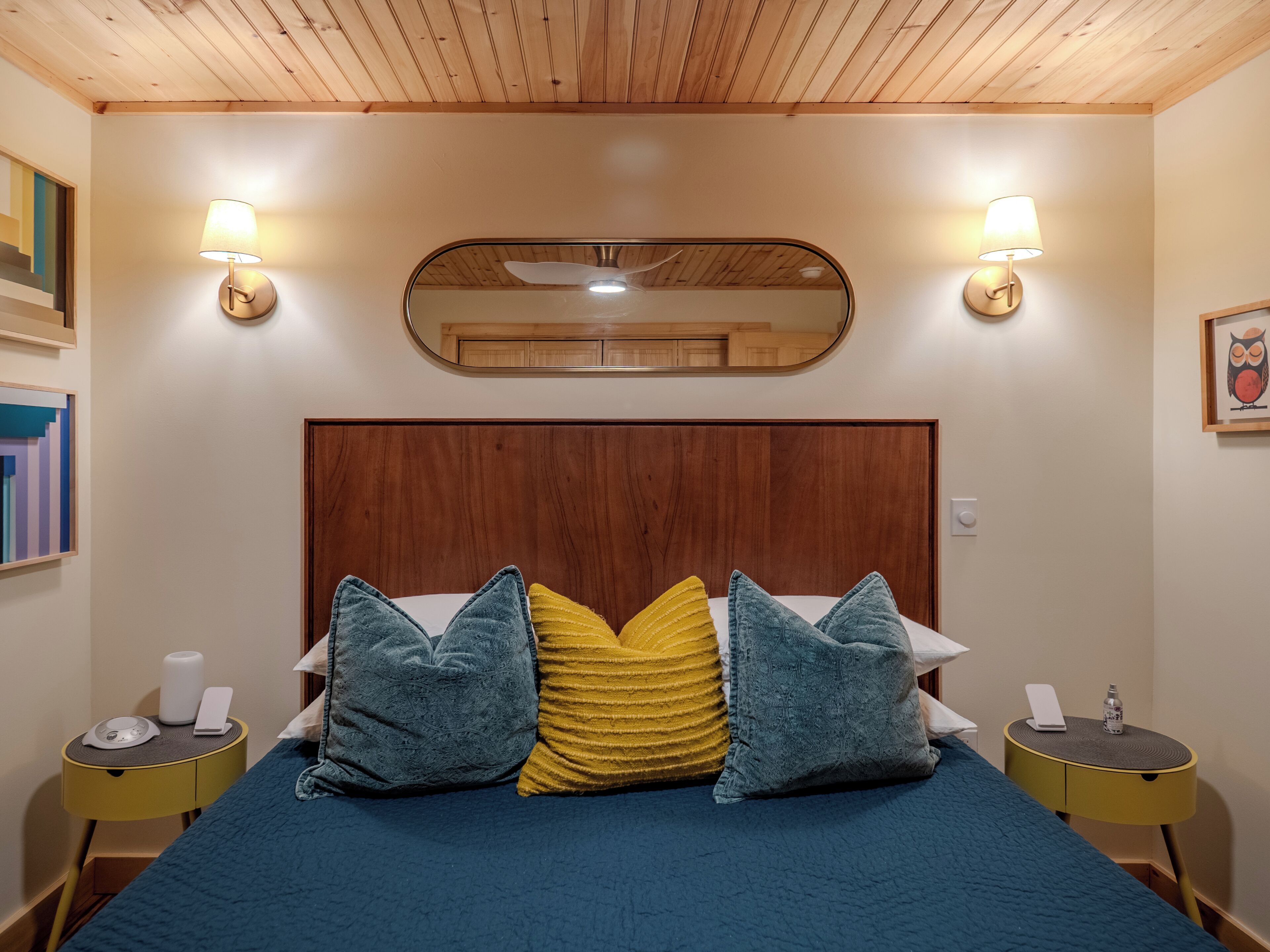 Habitación de hotel con techo de madera, cama grande con colcha azul, dos mesitas de noche con lámparas y un espejo sobre la cama.
