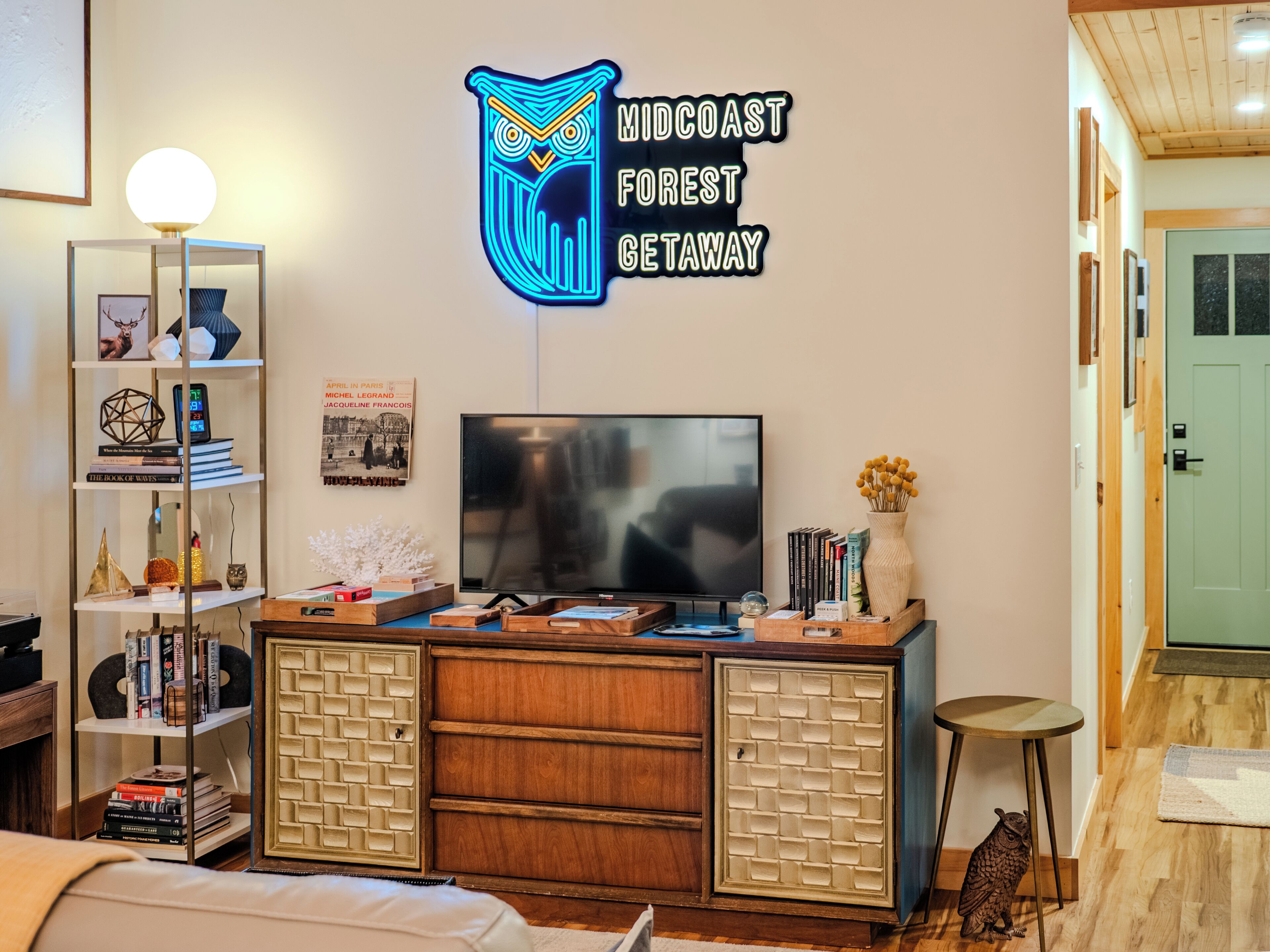 A modern living room with a flat-screen TV, a neon sign, a bookshelf, and a decorative owl statue.