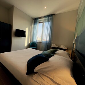 A hotel room with a large bed, a flat-screen TV mounted on the wall, a chair, a small table with a phone and a lamp, and a window with sheer curtains. - ELEMENTS#1 (Roma)