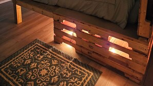 A bedroom with a bed on a wooden pallet base, a small bedside table with a lamp, a decorative rug, and a 'RELAX' sign on the wall.