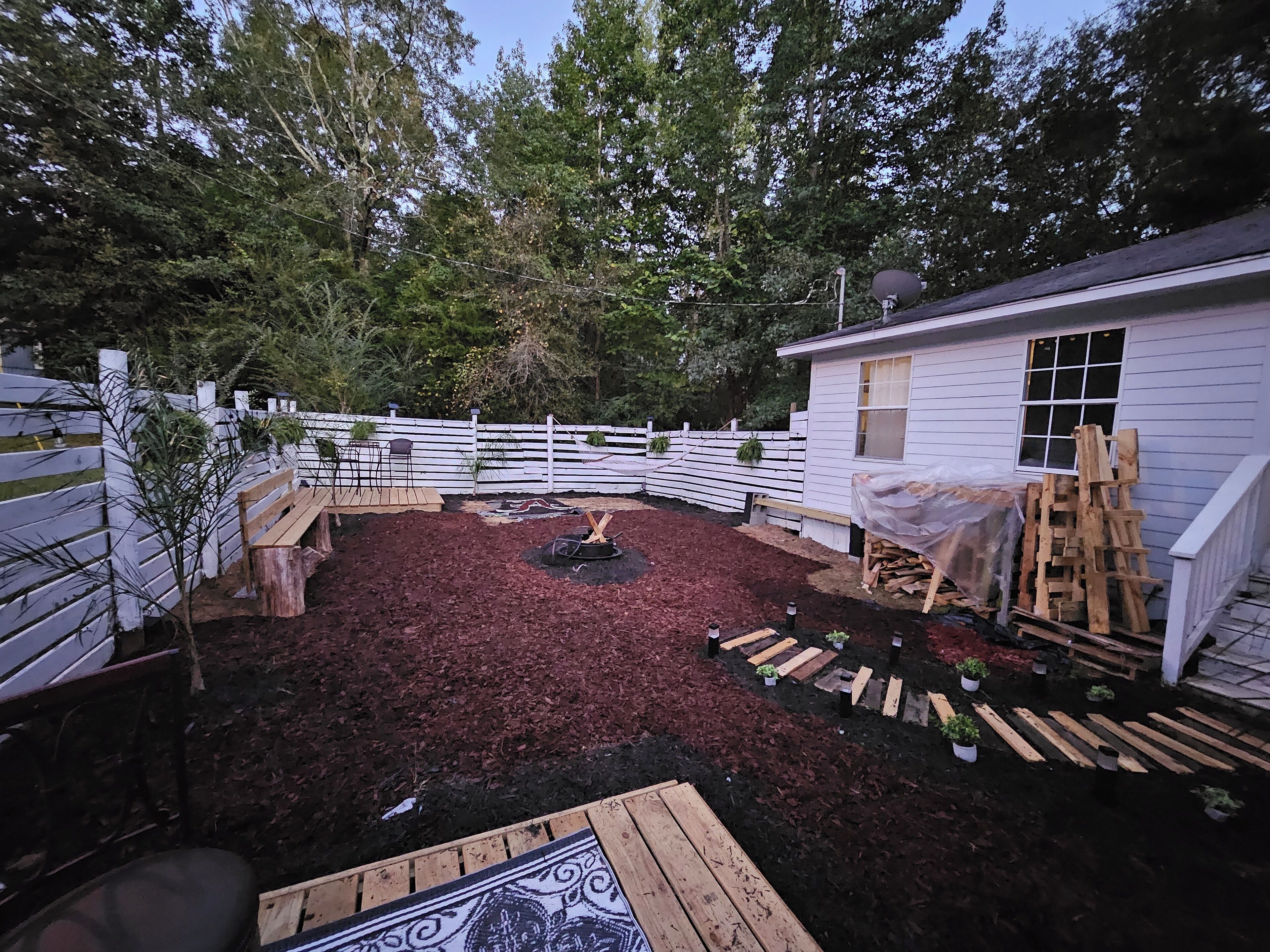 A fenced backyard with a wooden deck, a fire pit, and a white building with a window.