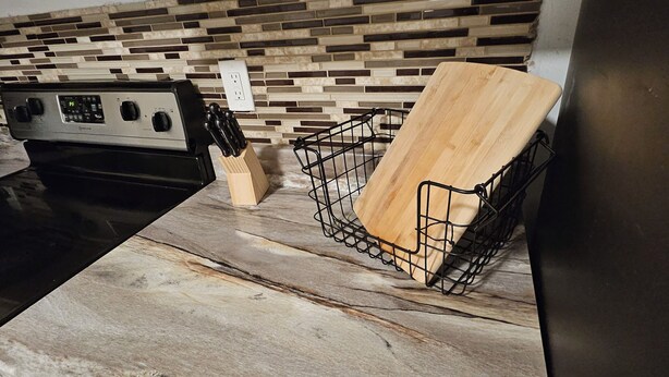 A kitchen with a granite countertop, a black wire basket, a wooden cutting board, and a stainless steel refrigerator.