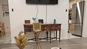 A modern living room with a round coffee table, a TV mounted on the wall, a dining area with a wooden table and chairs, and a kitchen with a stainless steel refrigerator.