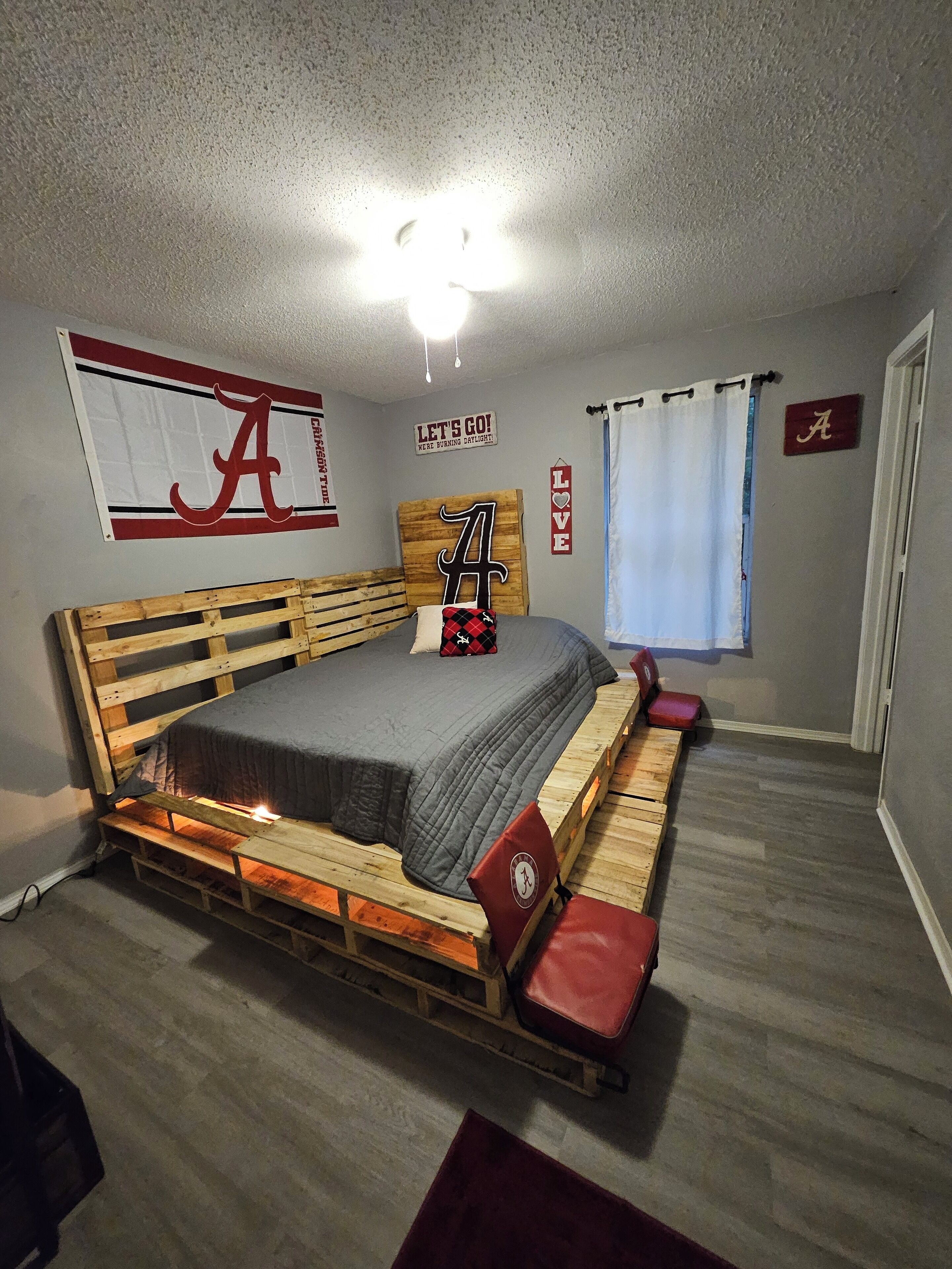 Ein Zimmer mit einem hölzernen Bett, einer Deckenleuchte, einer Wand mit einem großen 'A'-Schild und einem fenster mit Vorhängen.