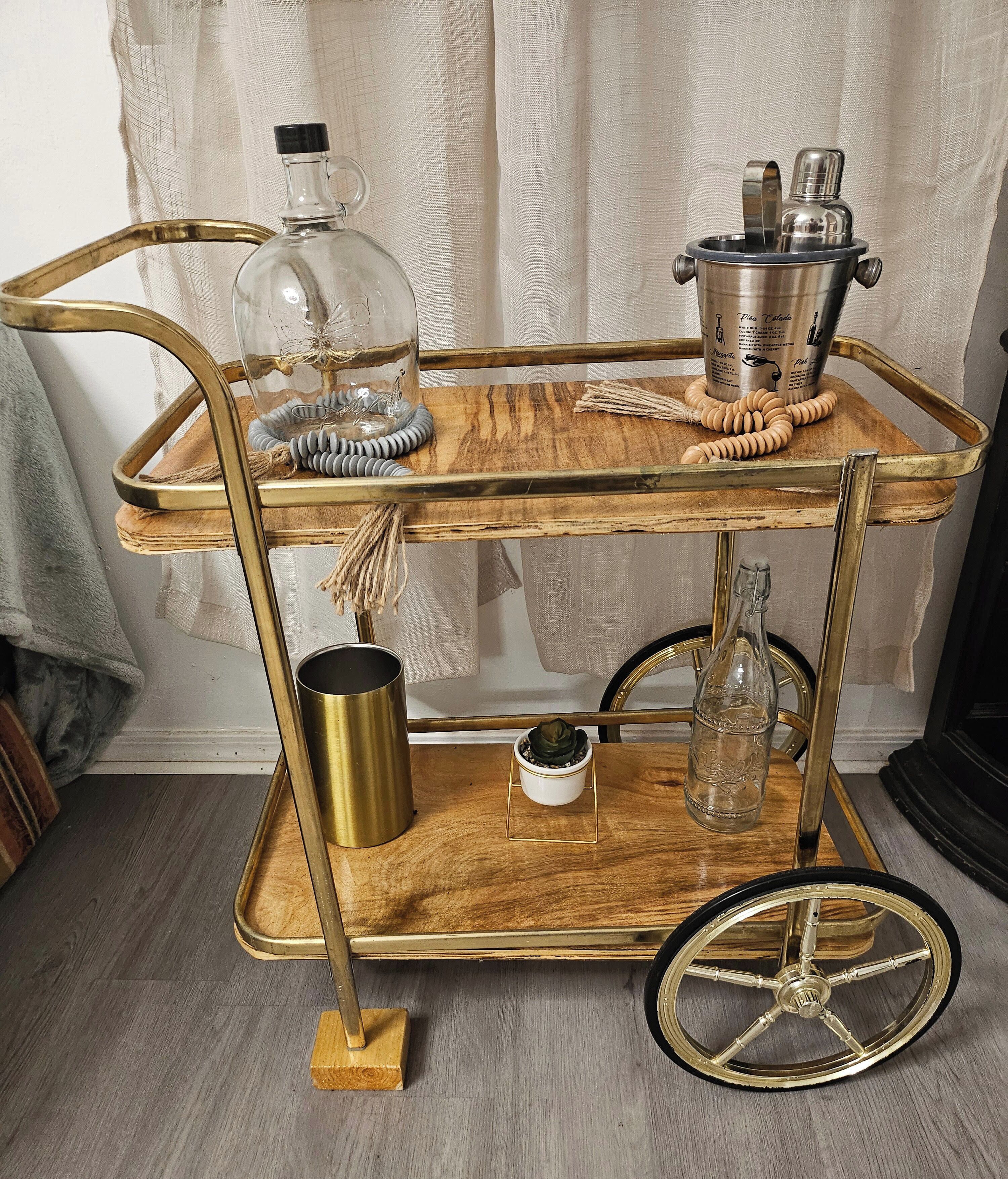 Carro metálico antiguo de dos niveles con tapa de madera, que incluye una campana de cristal, una jarra de metal y un pequeño cuenco blanco.