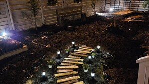 A nighttime outdoor area with a wooden pathway leading to a seating area, surrounded by plants and illuminated by small lights.