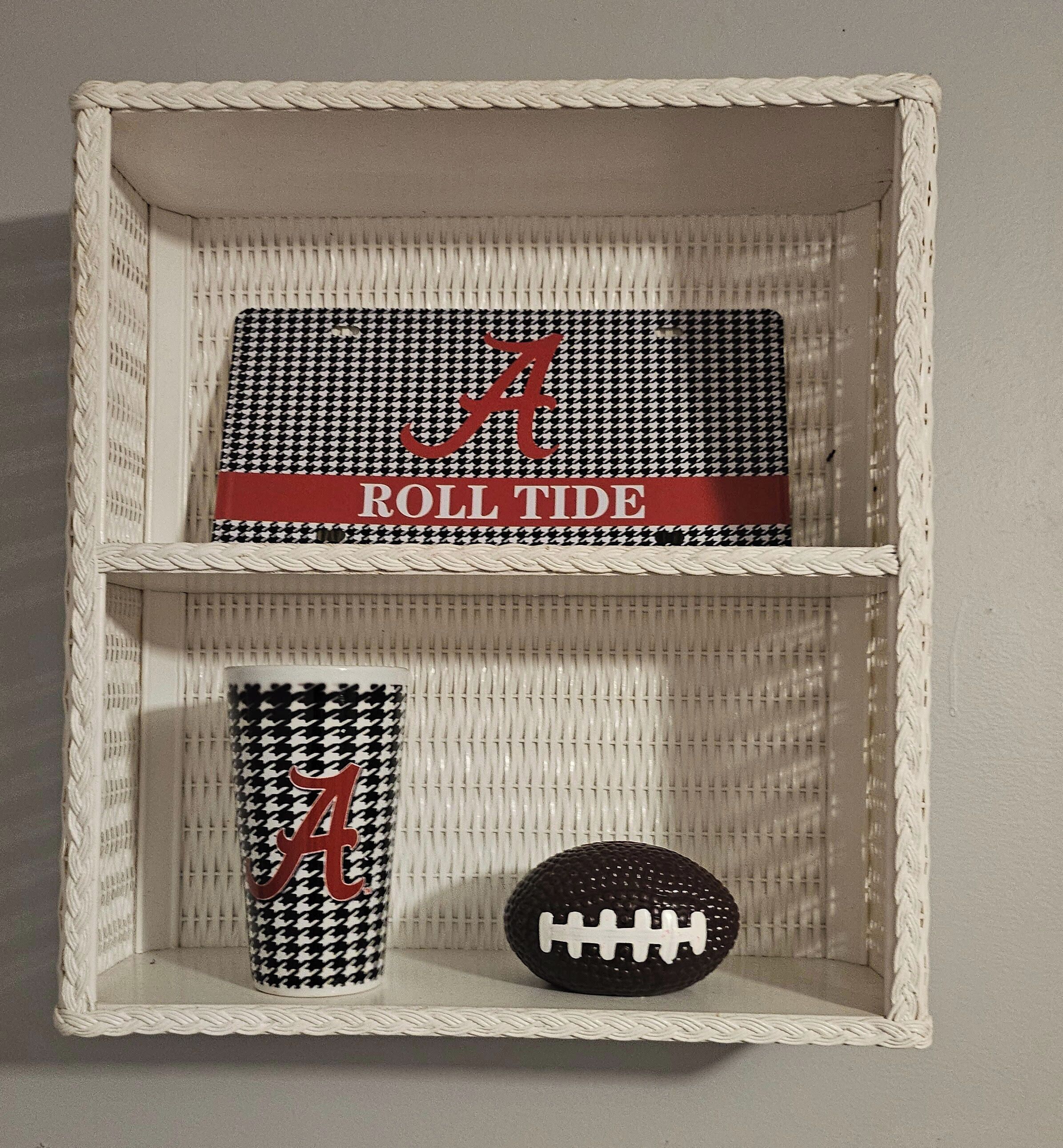 Una cesta tejida contiene un artículo con estampado pied de poule blanco y negro, que lleva el texto 'A' y 'ROLL TIDE' en rojo, un recipiente cilíndrico con el mismo estampado blanco y negro y una letra 'A' roja, y un balón de fútbol negro con cordones blancos.