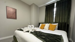Un lit bien fait, avec une couette blanche, un plaid vert foncé et deux oreillers jaune et blanc. Une serviette est disposée en forme de cœur sur le lit.