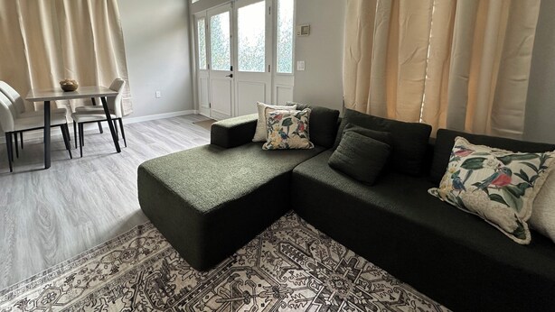 Un salon moderne avec un canapé d’angle, une table à manger avec des chaises et une grande fenêtre avec des rideaux.
