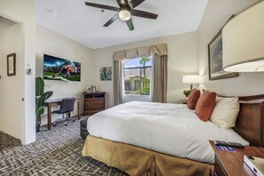 A hotel room with a large bed, a desk with a chair, a ceiling fan, a window with curtains, and a painting on the wall. - Desert Breeze: #244514 1 BR, communal pool! (La Quinta)