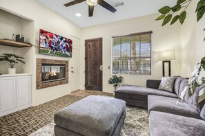 A living room with a fireplace, a television mounted on the wall, a sofa set, and a window with blinds. - Desert Breeze: #244514 1 BR, communal pool! (La Quinta)