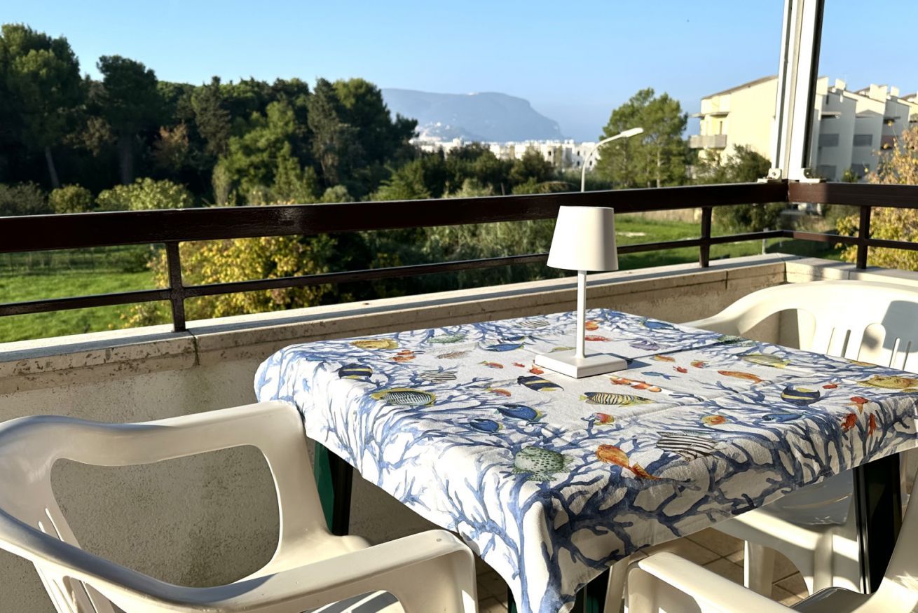 A balcony with a table covered in a fish-patterned tablecloth, white plastic chairs, and a lamp.