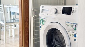 A front-loading washing machine with a control panel, a small table, and chairs in the background. - M410 - Marcelli, comfortable three-room apartment with large terrace (Marcelli)