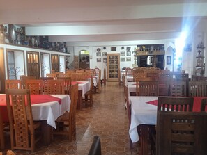 Una zona de menjador amb diverses taules i caires de fusta, un paviment de rajola i una cuina visible al fons.