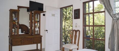 A bedroom with a wooden dresser, a television, a chair, a bed, and a window with a view of greenery.