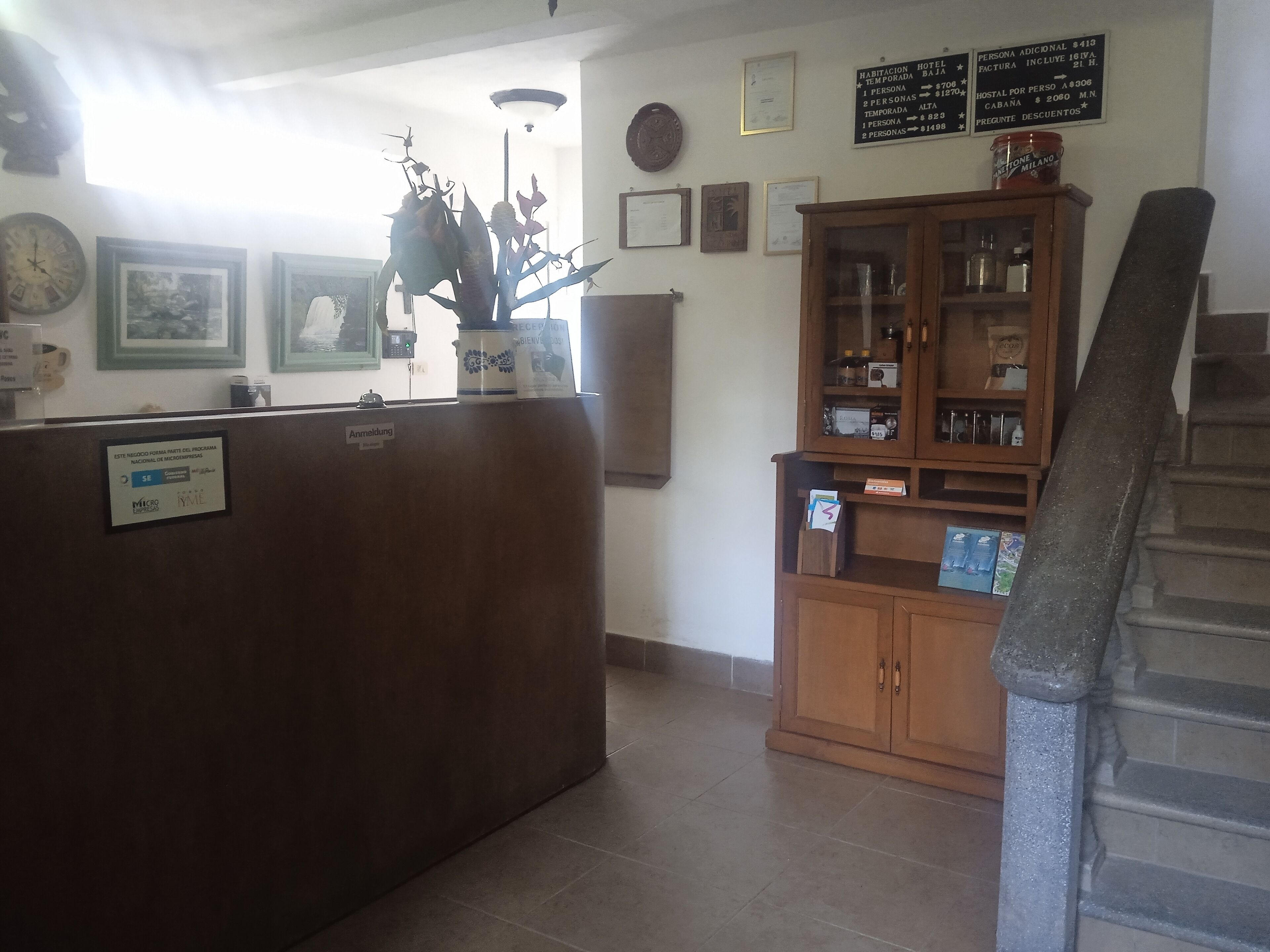 Un área de recepción con mostrador de madera, cuadros en la pared, un reloj y un cartel con tarifas de habitaciones.
